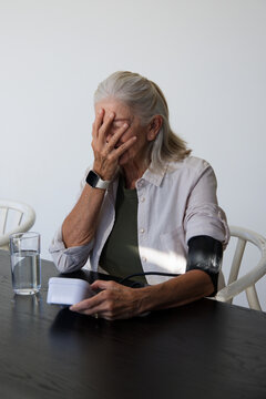 Senior female covering face while holding digital monitor and blood pressure cuff at home table