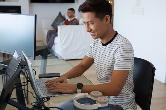Diverse male housemates working in living room, Asian man typing on laptop at glass desk