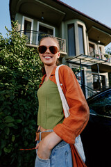 Obraz premium Smiling young woman in sunglasses and orange shirt poses near greenery and a house on a sunny day. Casual summer lifestyle, confident happy mood, street fashion portrait concept.