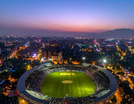 Aerial view of a brightly floodlit cricket stadium packed with spectators during a dusk match against a sprawling city skyline. Ideal for sports entertainment and urban leisure concepts.