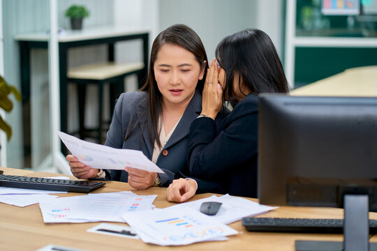 Asian businesswomen whispering confidential information and secret office gossip at desk during working hours. Corporate women discussing financial paper report.
