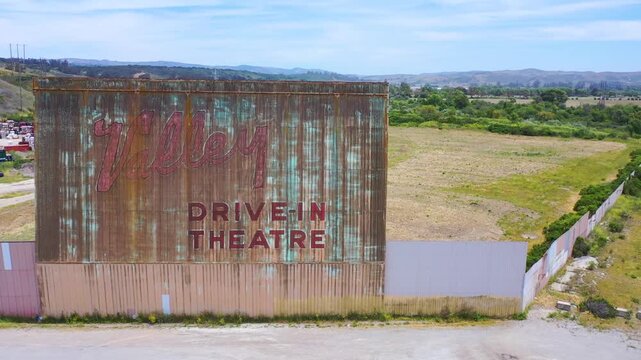 Aerial view showing an abandoned drive-in movie theater with its weathered screen and empty lot, slowly revealing the back of the screen and a highway with moving cars in the distance.