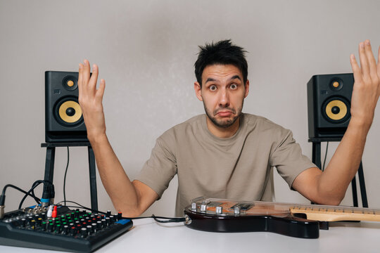 Confused man shrugging and raising hands in question, encountering technical difficulties with home music studio setup, audio mixer and bass guitar on white table, isolated background, copy space.