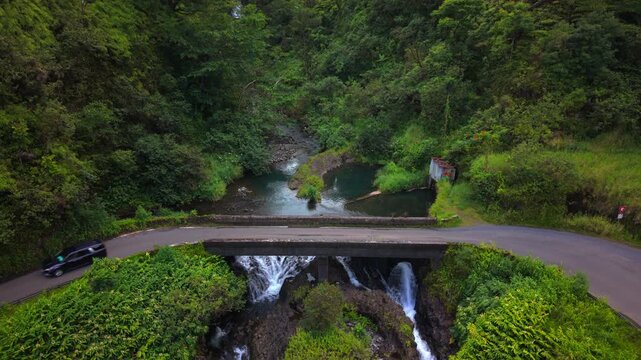 Aerial view of dark van crossing narrow concrete bridge above cascading waterfall on the road to Hanna on Maui Hawaii island USA surrounded by lush tropical rainforest and a green river gorge.