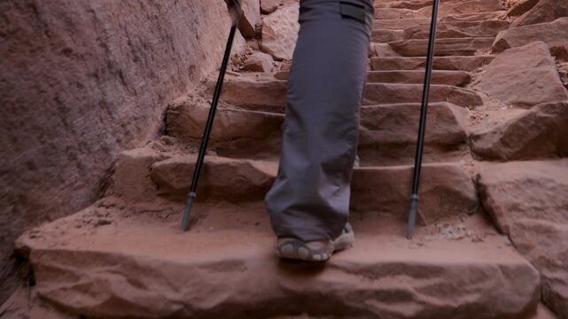 Close up view of trekker feet and legs rises upward steep, rugged stone staircase carved into red rock canyon, using trekking poles for stability during an adventurous hike