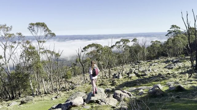 A girl hiker stands and admires the thick clouds emerging above the eucalyptus bush and a meadow, from the summit of Mount Macedon, Macedon Ranges, near Melbourne, Victoria, Australia