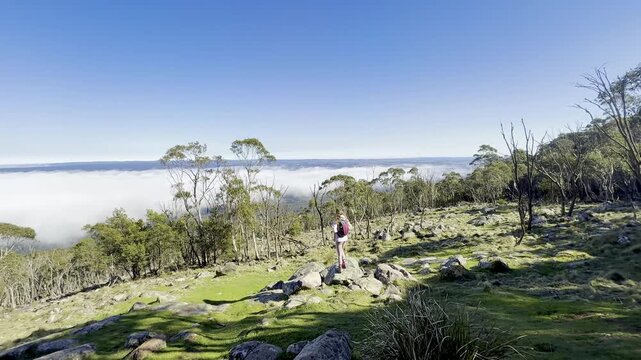 A girl hiker stands and admires the thick clouds emerging above the eucalyptus bush and a meadow, from the summit of Mount Macedon, Macedon Ranges, near Melbourne, Victoria, Australia