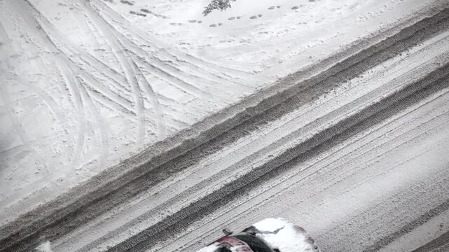 Aerial view of car driving on snowy road during winter storm in city leaving tire tracks on ice