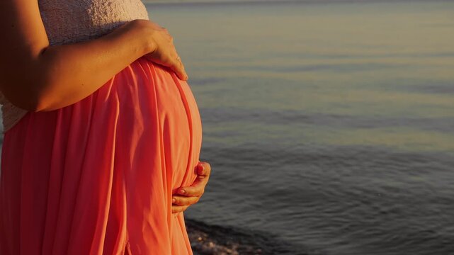 Close-up of a pregnant woman gently holding her baby bump by the water in warm sunset light, symbolizing maternity, motherhood, prenatal care and anticipation of a baby.