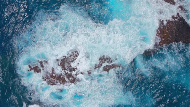 Aerial top down view of Kaihalulu Beach on Maui Hawaii island where turquoise waves and white foam swirl around volcanic black rocks in deep blue ocean water along rugged tropical coast.