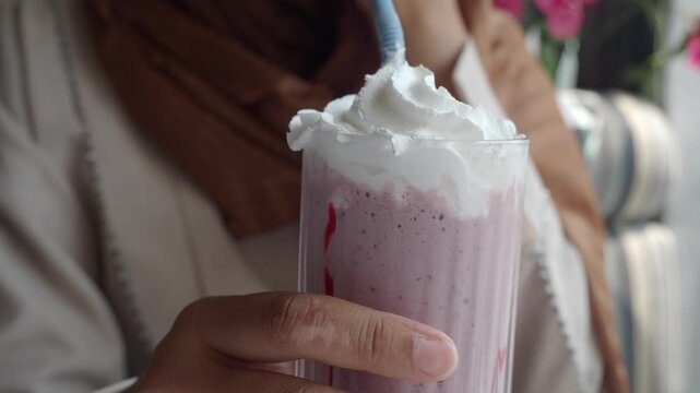 Close up shot of woman in hijab drinking strawberry milkshake through straw with whipped cream