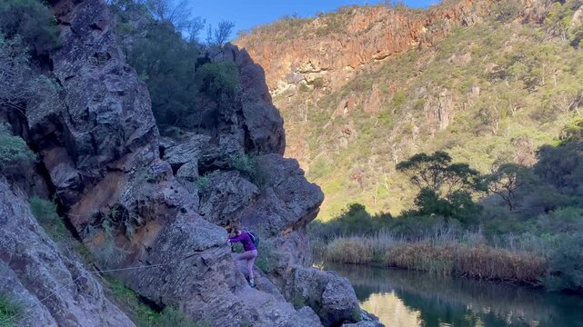 A girl hiker rock scrambles along a cliffside, holding the cable handrail above the river, on a tough section of the Werribee Gorge Circuit Walk, near Melbourne, Victoria, Australia.