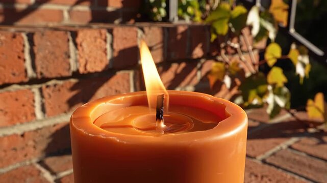Steady golden flame rising from dark wick inside a weathered orange pillar candle on red brick, backed by softly blurred climbing foliage for an inviting autumn patio atmosphere and seasonal comfort