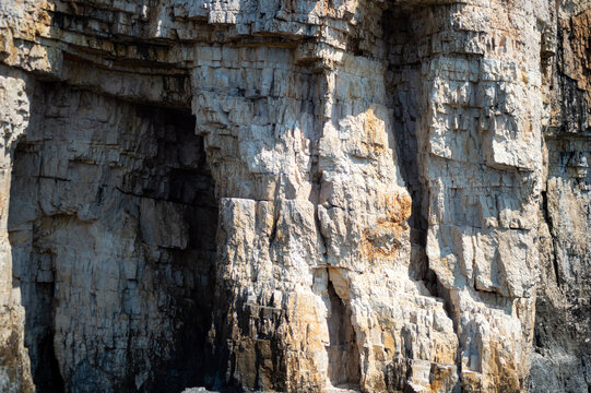 A close-up view shows rugged limestone cliffs along the sea coast in Corfu, Greece. Layered stone formations and natural rock alcoves form the coastal cliff face under daylight.