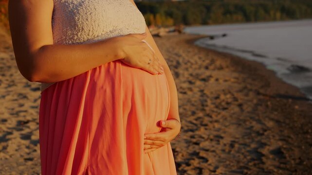 Close-up of a pregnant woman gently holding her baby bump on a sandy beach in warm sunset light. Peaceful maternity scene symbolizing motherhood, care and anticipation.