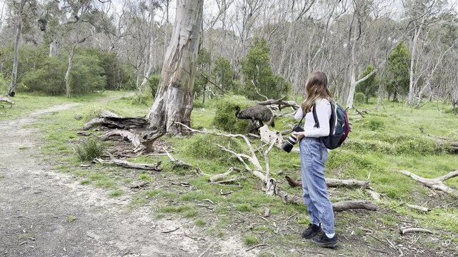 A happy girl photographer with a camera smiles while standing very close to an emu at Nangak Tamboree Wildlife Sanctuary, La Trobe University, Bundoora, Melbourne, Victoria, Australia.