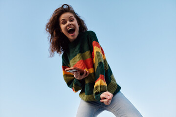 Excited young woman with curly hair in colorful striped sweater shouting with joy while holding...