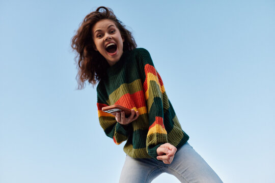 Excited young woman with curly hair in colorful striped sweater shouting with joy while holding smartphone against blue sky. Concept of happiness, success and freedom.