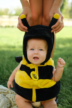 A mother holds a baby dressed in a bumblebee costume, sitting on the green grass in a park; a childhood theme.