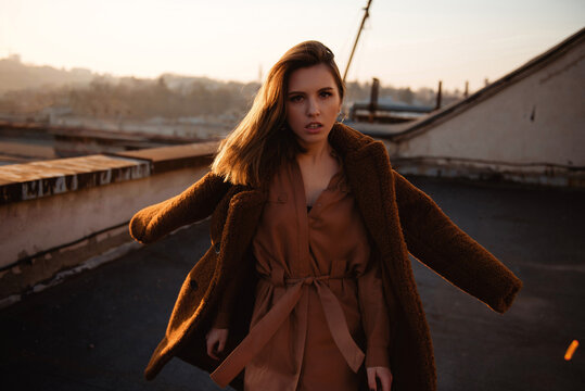 Woman in brown coat and dress on rooftop at sunset