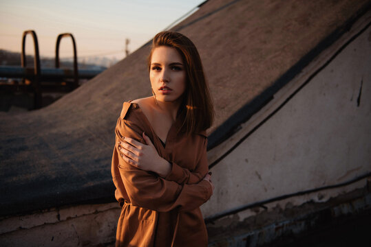 Young woman embracing herself against a backdrop of industrial structures at sunset
