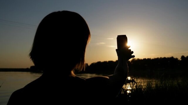 Backlit silhouette of a person raising a beverage can against the setting sun by the water. Warm evening light outlines the subject, offering an evocative tone for summer drink promotions.
