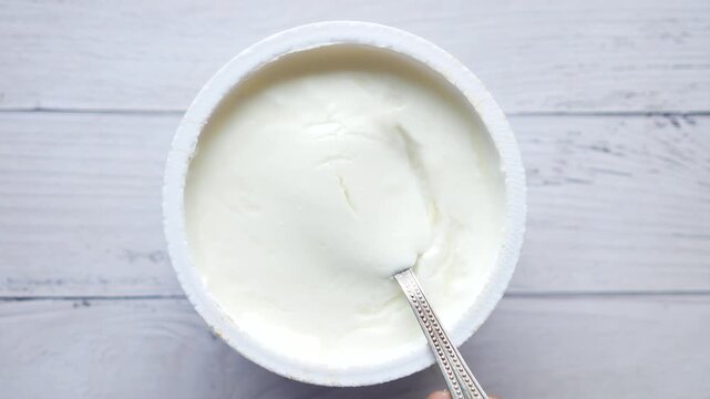 Close up top view of a spoon scooping creamy yogurt from a white plastic container, dynamic motion clip