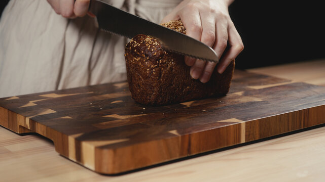 Close-up of baker in rustic linen apron slicing freshly baked artisan whole-grain seed bread with knife on wooden cutting board. Homemade loaf for breakfast, bakery promos, and healthy food concepts.