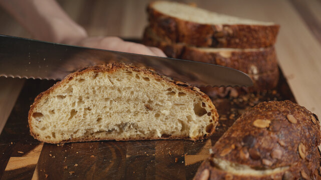 Close-up of hands slicing freshly baked artisan sourdough bread with a knife on a wooden cutting board. Homemade crusty open crumb loaf for breakfast, bakery promos, and healthy food concepts.