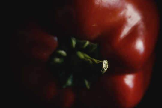 Macro shot of ripe red bell pepper with vibrant green stem in moody lighting on dark background. Fresh raw vegetable for organic recipes, culinary menus, nutrition, and healthy eating.