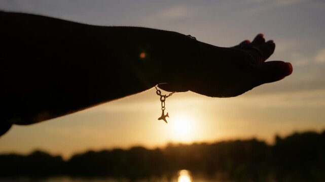 A woman extends her hand wearing an airplane charm bracelet against a bright sunset. Warm rim lighting offers an evocative wanderlust visual for travel promotions.