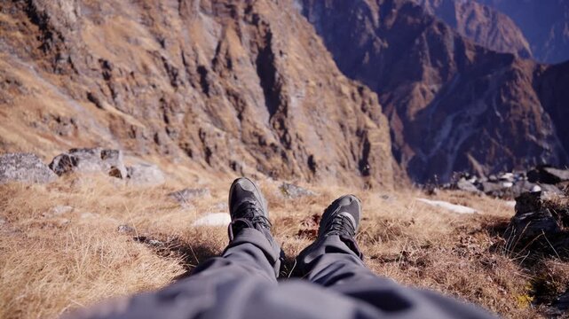 POV shot of a trekker's two hiking boots stretched out on golden dry grass with dramatic rocky mountain cliffs and patchy snow in background