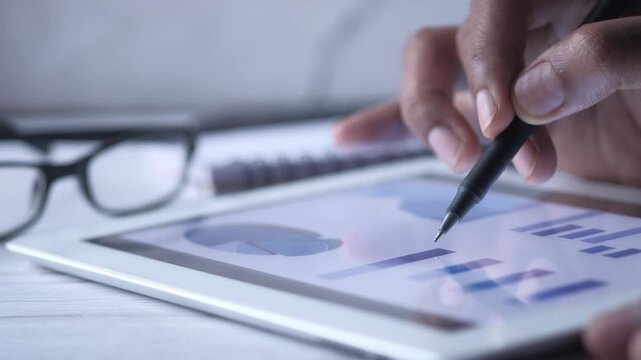 Close-up of hand using stylus on tablet with financial charts, glasses and notebook in background