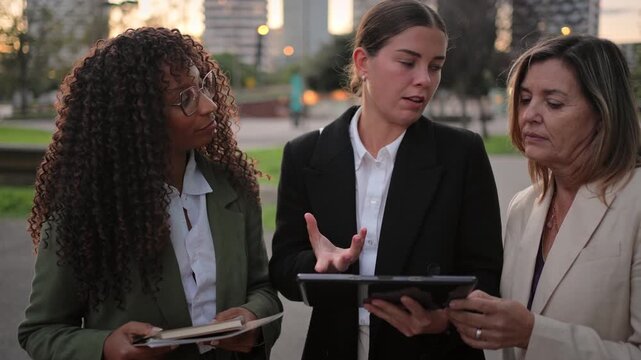 Three business women coworkers discussing project on tablet standing in modern in corporate city district