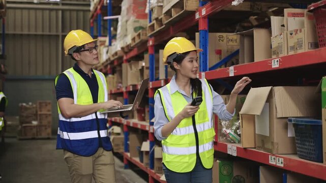 Team of Asian warehouse workers wearing yellow safety helmets holding and using a laptop and a barcode scanner to check inventory stock on shelves together in a logistics distribution factory center. 