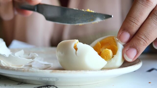 Close up shot of person using knife to cut hard boiled egg on plate with shell pieces