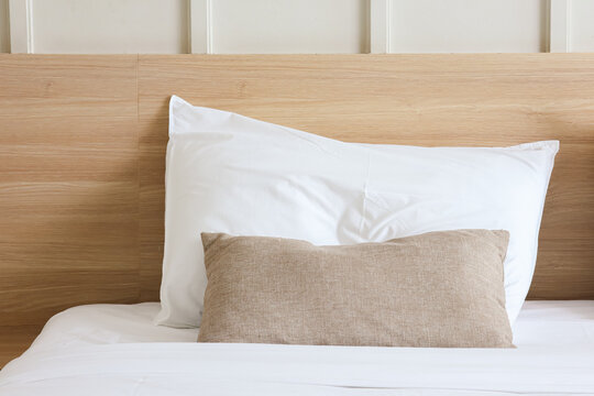 Close-up of fluffy white and brown decorative pillows neatly arranged on a white bed sheet against a modern wooden headboard in a cozy bedroom