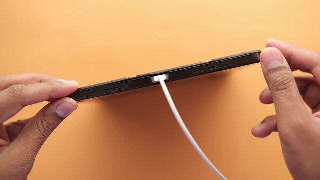 Hands plugging usb type c white cable into tablet charging port on orange background