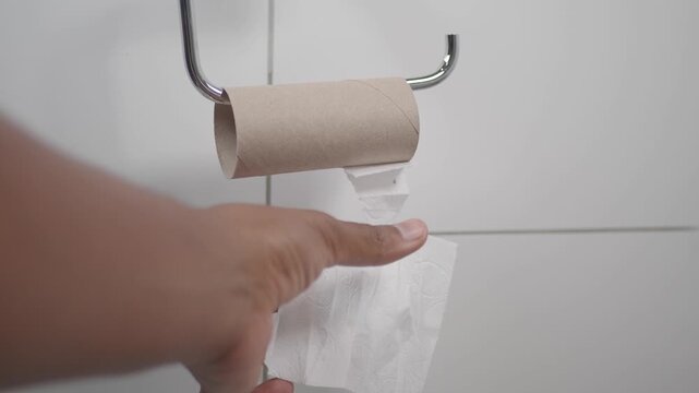 Person tearing off last piece of paper from empty toilet roll in bathroom over white tiles background