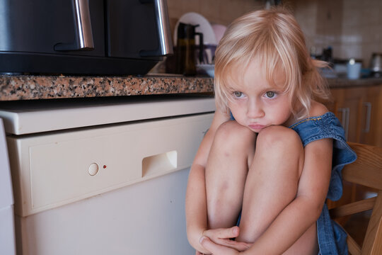 Offended little blonde girl sitting curled up on chair in kitchen