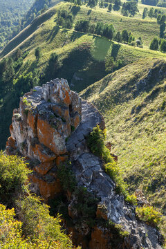 A rugged lichen-covered rock formation juts from a sunlit hillside, overlooking rolling green valleys and scattered trees. Serene mountain landscape evokes adventure, solitude and nature exploration