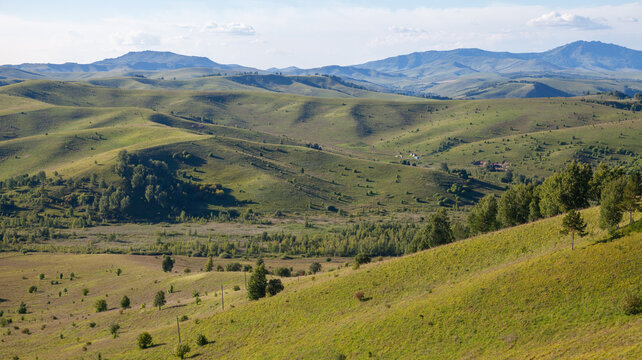 Expansive panoramic landscape of rolling green hills and pastoral countryside under a blue sky, with scattered trees, sunlit valleys and distant mountains conveying calm, openness and rural serenity