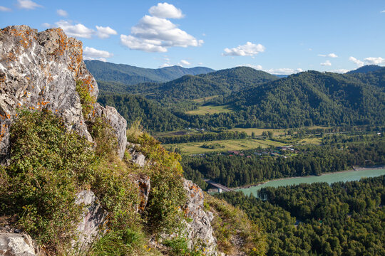 A panoramic scenic landscape showing a rocky cliff overlooking a green valley, winding river and forested mountains under a bright blue sky with scattered clouds, evoking peace and adventure