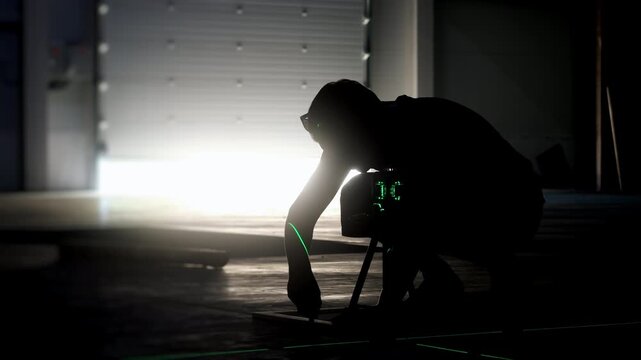 Worker uses a green laser level for precise measuring and alignment on the floor inside an industrial building, with strong backlight from an open doorway.