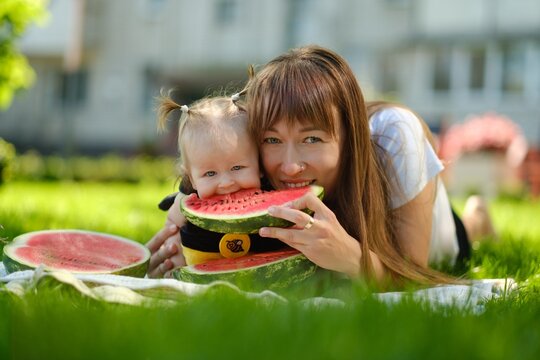 Happy mother and baby girl eating fresh red watermelon slice on green grass outdoors picnic, family summer concept.