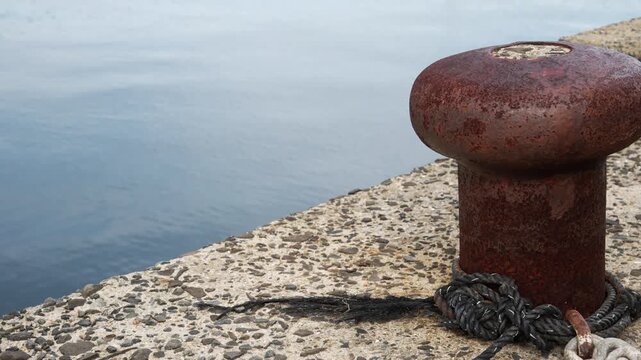 4K cinematic footage of a rusty mooring bollard with rope on a concrete pier by the calm sea