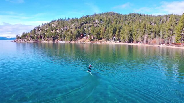 Person riding hydrofoil board across clear blue lake water with mountain forest scenery.