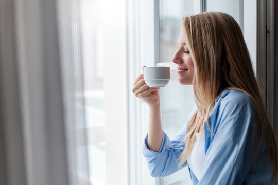 Young woman enjoying coffee, morning routine, calming drink at home. Happy girl relaxing. Concept of slow living, mindfulness, female mental health. Fresh air, wind from opened window