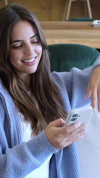 Young happy brunette woman relaxing at home on the sofa while texting, browsing social media or shopping online on her smartphone, enjoying a peaceful moment of digital connection