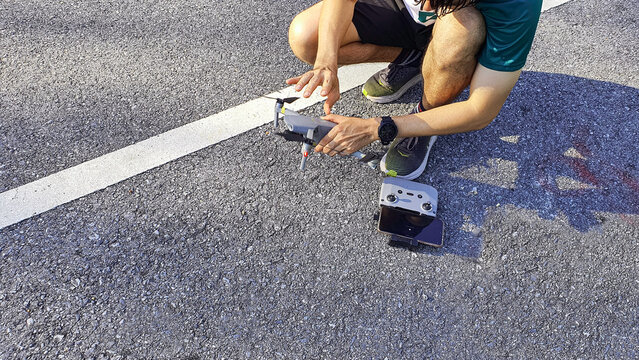 A person preparing and operating a compact camera drone on an asphalt road, with a remote controller and smartphone. Outdoor technology concept representing aerial photography